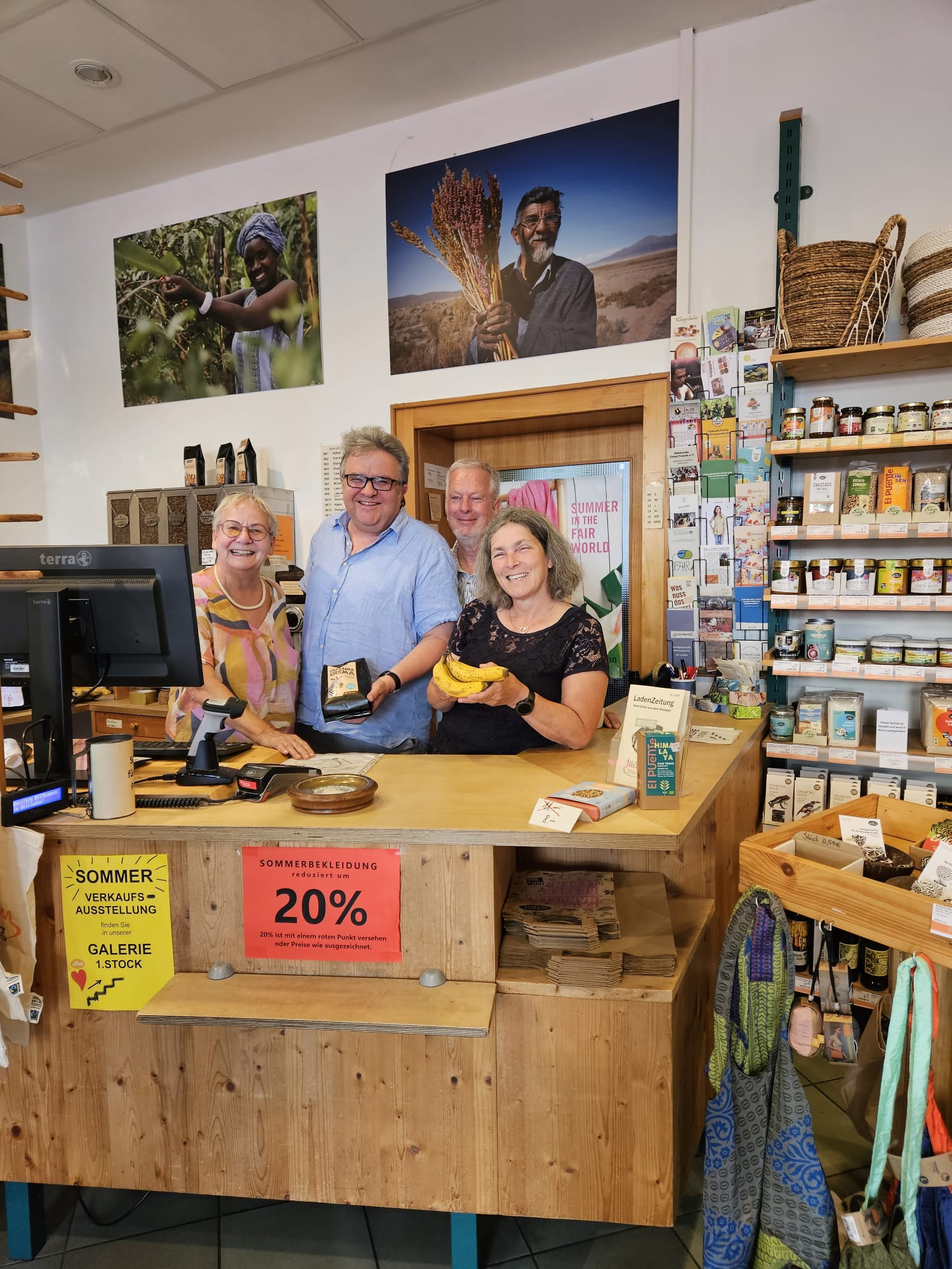 Zu sehen sind Kerstin Celina, Gerhard Müller und zwei Mitarbeitende aus dem Eine-Welt-Laden hinter einer Ladentheke. Kerstin Celina hält Bananen in der Hand, Gerhard Müller eine Packung Kaffee. Alle vier Personen lächeln in die Kamera